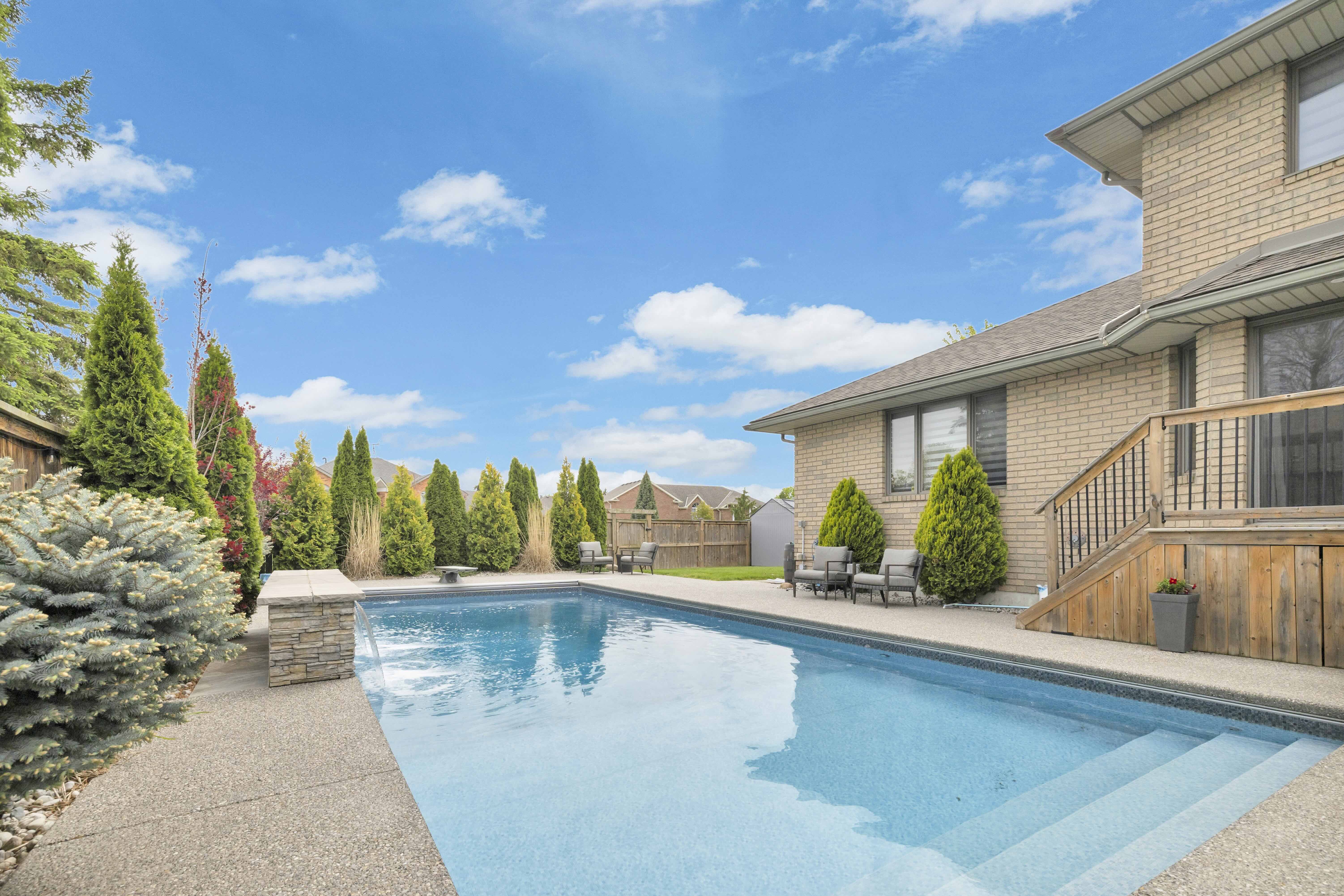 Inground pool with stone column, wooden deck and privacy trees