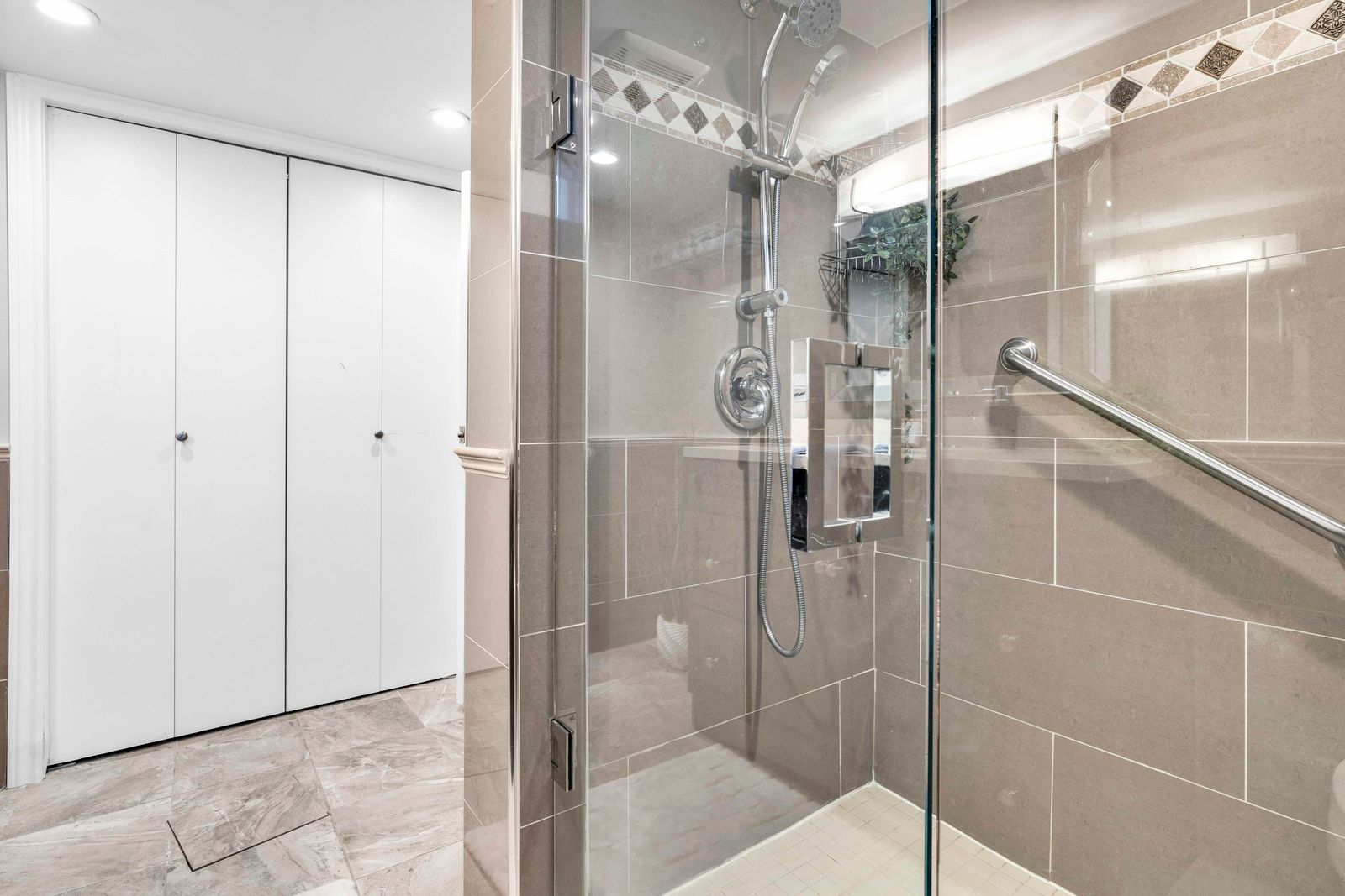 Glass-enclosed shower with decorative tiles and grab bar next to a closet with white doors in a bathroom.