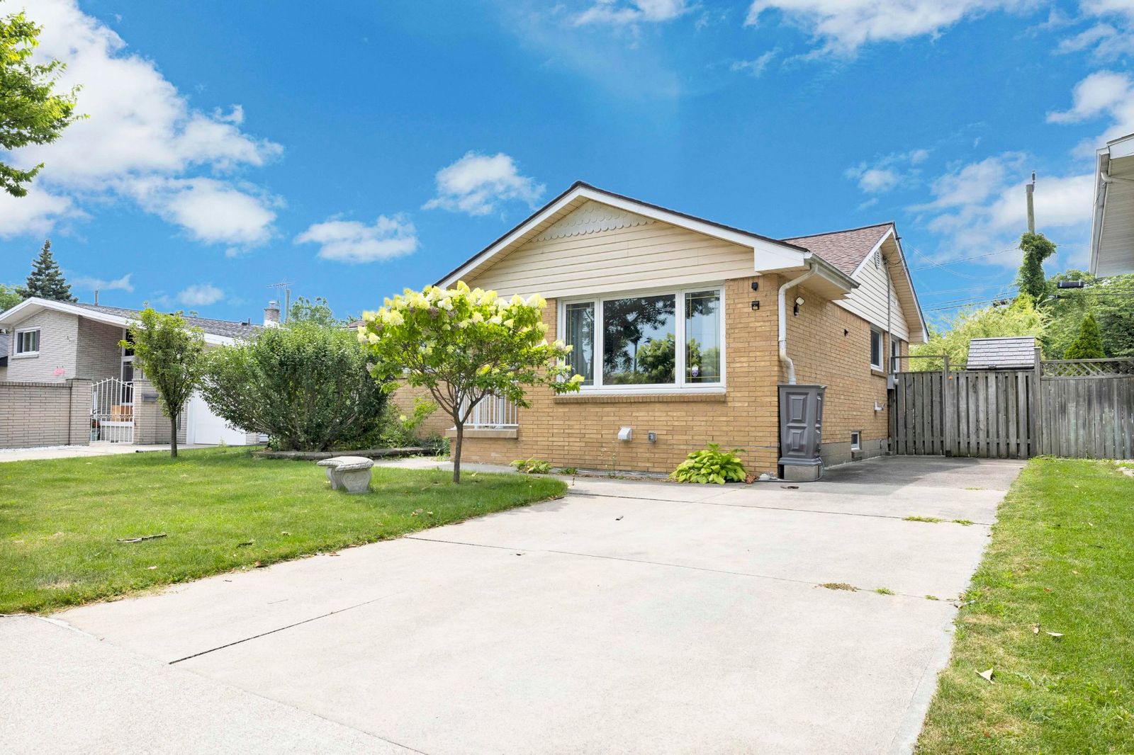 Side exterior view of a brick ranch house with a large window, concrete driveway, and green lawn.