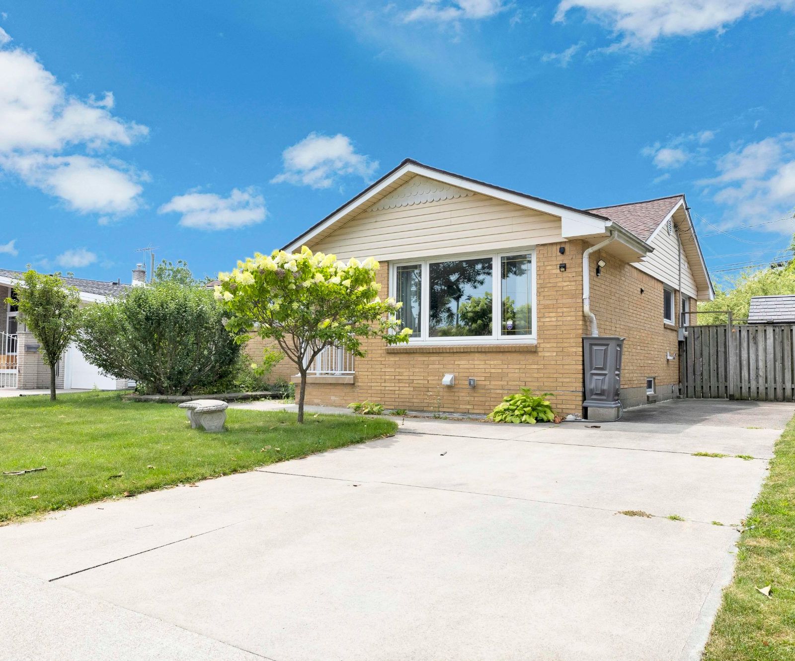 Side exterior view of a brick ranch house with a large window, concrete driveway, and green lawn.