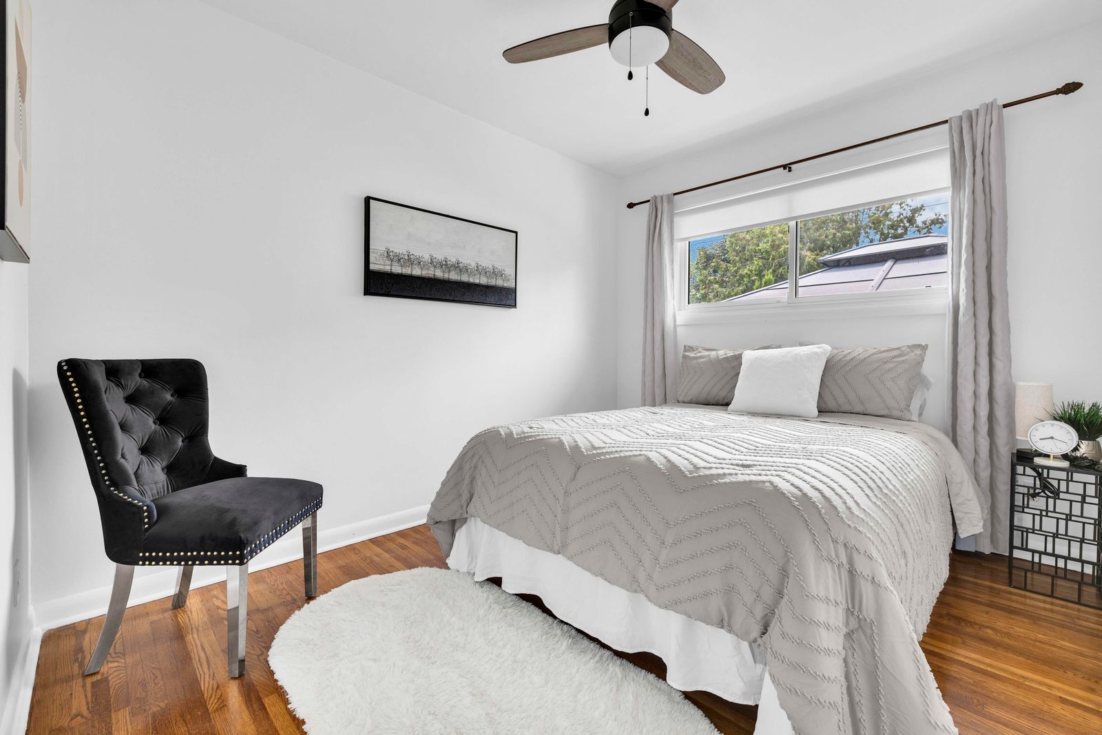 Bright bedroom with a bed, black tufted chair, white walls, and a ceiling fan.