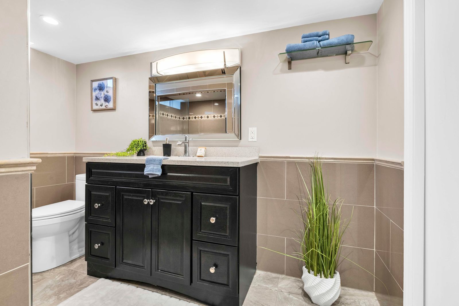 Bathroom vanity with framed mirror, toilet, and decorative plants with tiled walls and floor.
