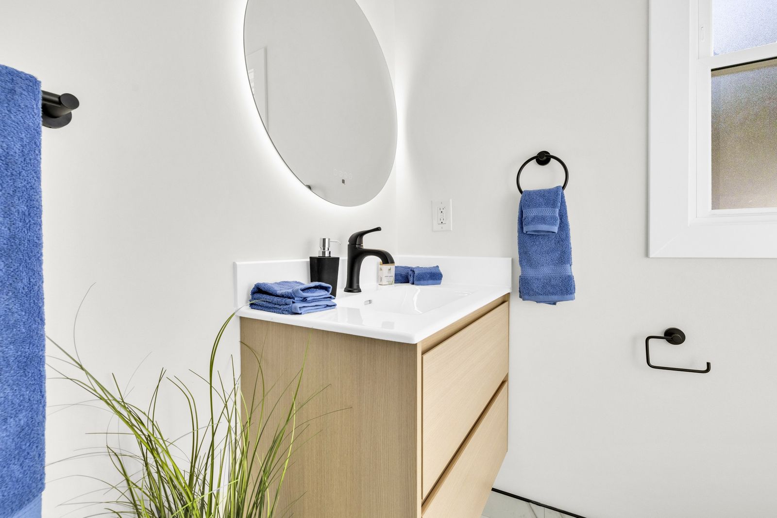 Bathroom vanity with a wood-tone floating cabinet, white top, black faucet and a round backlit mirror.