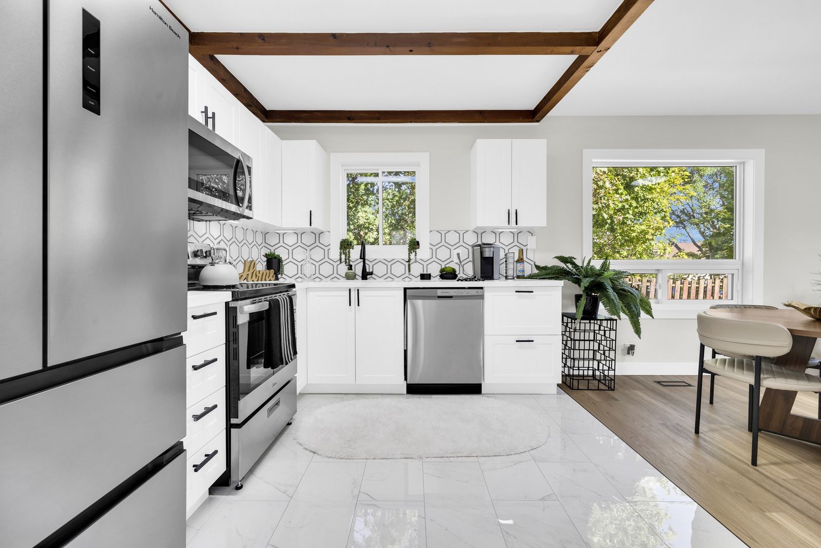 Kitchen with white cabinets and black hardware, stainless steel appliances, hexagon backsplash and two windows, beside the dining table.