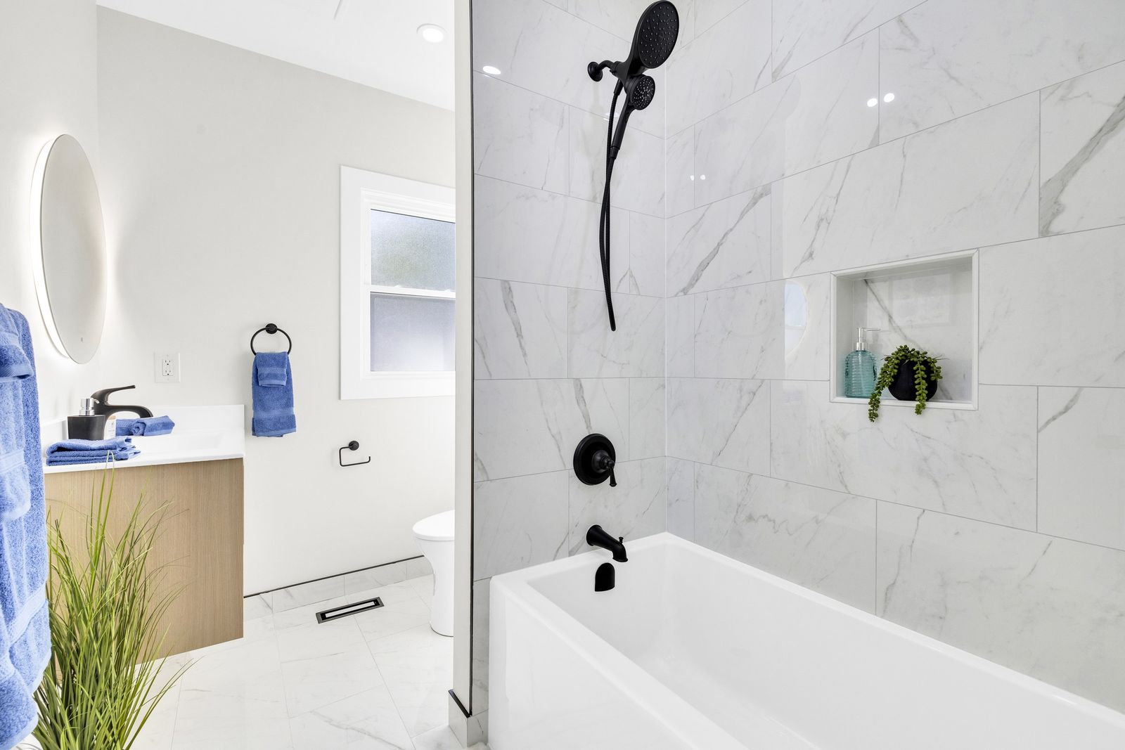 Bathroom with a bathtub in a tiled surround, black shower fixtures, a vanity and a round mirror.