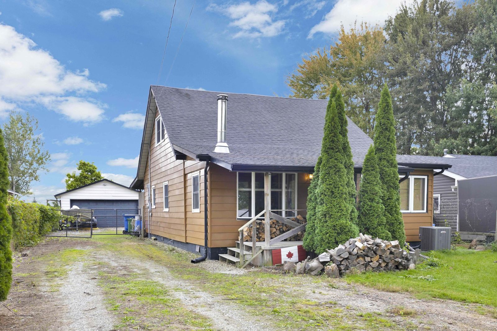 Side of the house with brown siding, evergreens, a gravel driveway and the detached garage behind.