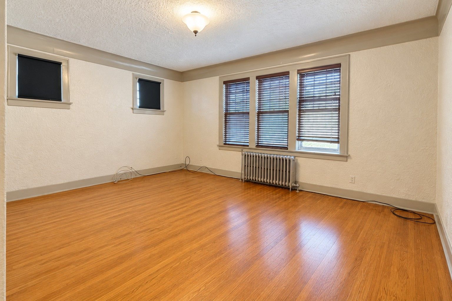 Empty living room with a wood floor, three windows along one wall and a radiator beneath them.