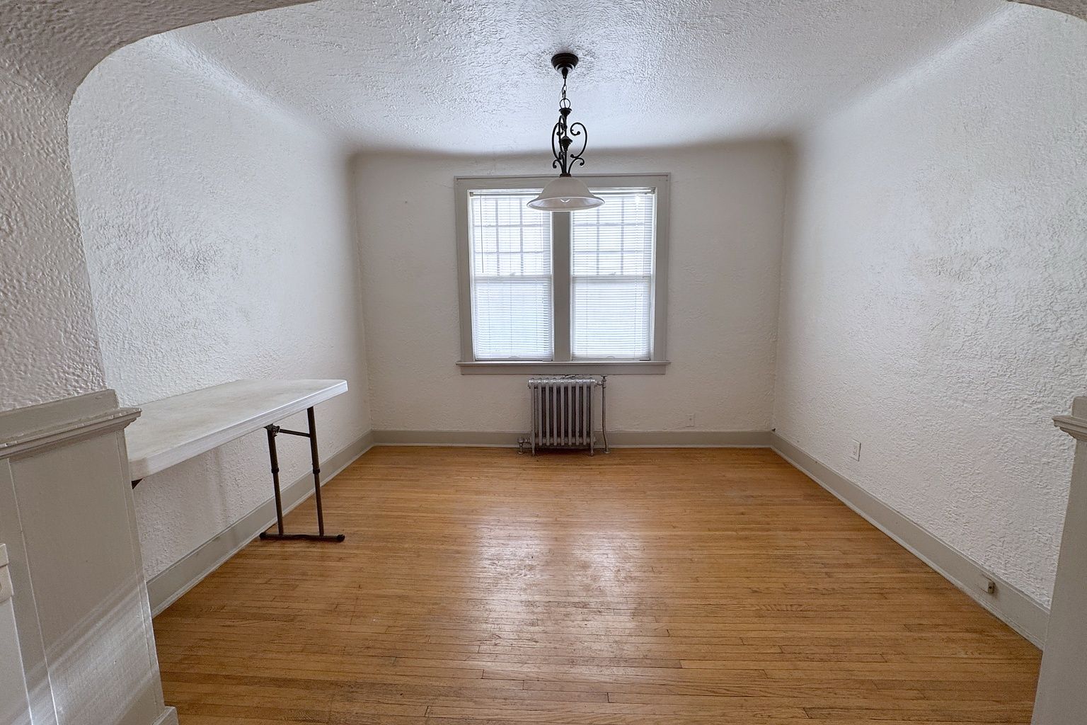 Empty dining room with a wood floor, a window with a radiator beneath it and a hanging light fixture.